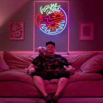 A fat, gender non-conforming person sits on a couch in a neon pink-washed living room. They hold stuffed animal pigs, and seem peaceful.
