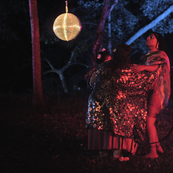 Three people in sparkly clothes sway together in dark, neon-lit woods under a disco ball.