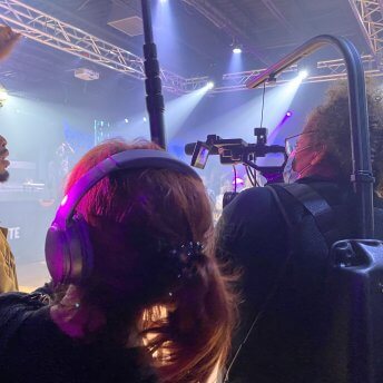 A curly-haired cinematographer and a red-headed sound recordist film at a nightlife venue. A young Black man cheers a performer on from the sidelines.