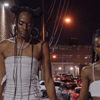 Two Black nonbinary femmes with long braids wearing white stylish outfits walk fabulously down a street at night.
