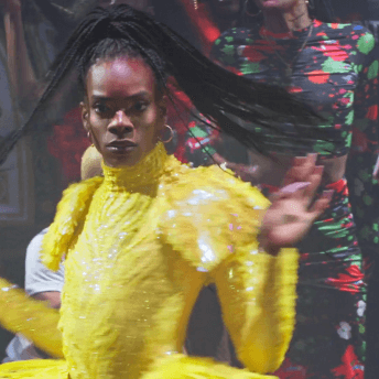 A Black nonbinary femme twirls on a ballroom floor wearing a yellow sequined dress, flipping her braids.