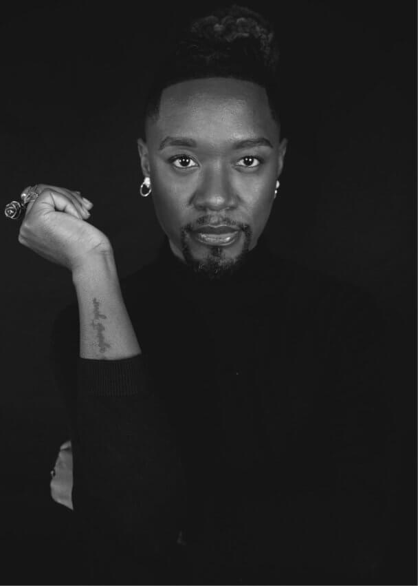 A Black nonbinary person with a goatee and earrings stares fiercely into the camera, with their hand by their face.