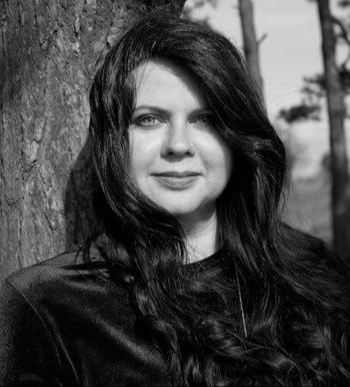 A white woman with long dark hair stands in front of a tree wearing a blue velvet shirt. Lindsey Dryden headshot in black and white.