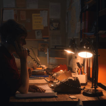 In a dimly-lit, messy office, two people sit at a desk speaking on rotary phones.
