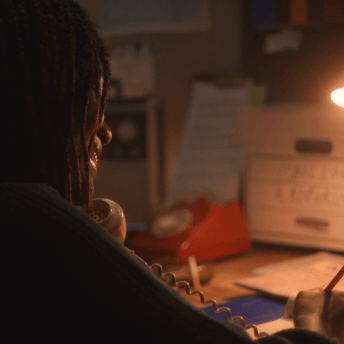 In a dimly-lit phone room, a Black woman with long braids speak on a rotary phone, smiling and writing on a note pad.