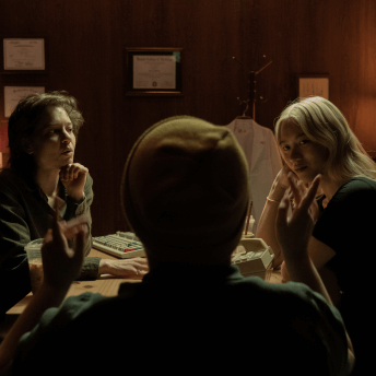 In a dimly-lit set that resembles a doctor's office, two actors look intently at the director gesturing to them.