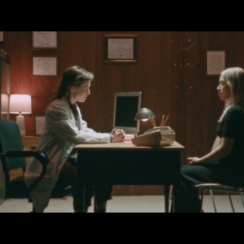 In a dimly-lit set that resembles a doctor's office, an actor dressed as a doctor leans menacingly across the table towards a patient.