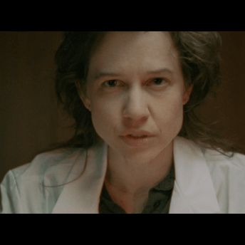 A nonbinary white person wearing a doctor's coat looks judgmentally at the camera in close up.