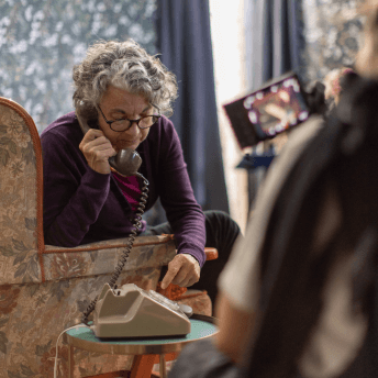 On a film set that looks like a living room, an older woman dials a rotary phone. The camera person is in the foreground out of focus.